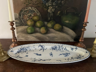 A vintage French large oval platter with a blue and white floral pattern displayed on a table with a painting of fruits in the background.